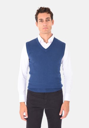 Maglione senza maniche lavorato a maglia blu con design a scollo a V, messo in risalto da una texture liscia e orlo a costine, indossato sopra una camicia bianca con colletto.