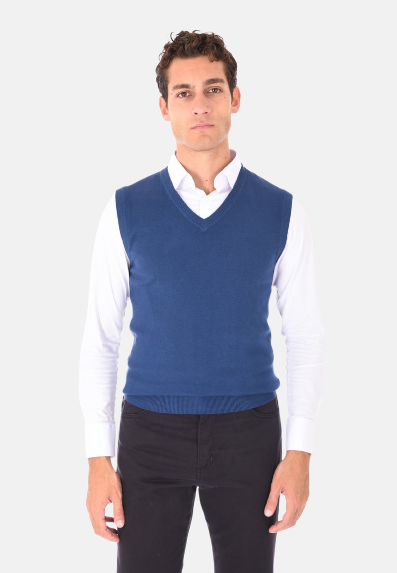 Maglione senza maniche lavorato a maglia blu con design a scollo a V, messo in risalto da una texture liscia e orlo a costine, indossato sopra una camicia bianca con colletto.