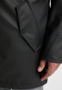 Close-up of a hand holding the collar of a sleek black jacket, featuring a visible pocket and button detail, with a neutral background.