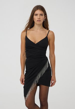 Abito bodycon nero con spalline sottili, caratterizzato da un design drappeggiato e una frangia di perline argentate lungo l'orlo su un lato.