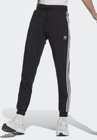 Pantaloni sportivi neri con strisce bianche sui lati, vita con coulisse e polsini a costine. Presenta un logo adidas sulla coscia sinistra.