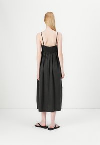 Abito maxi in lino nero con corpetto arricciato, spalline sottili e gonna ampia e fluida. Sandali indossati; sfondo semplice.
