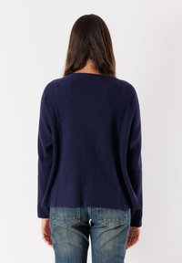 Donna con lunghi capelli castani, che indossa un maglione a coste blu navy e jeans blu, in piedi di fronte a uno sfondo bianco.