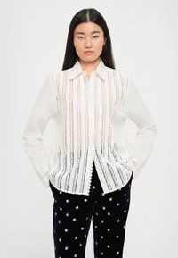 Balta marškiniai su ilgomis rankovėmis, horizontaliais nėrinių elementais, klasikiniu apykaklės dizainu ir permatomu, tekstūruotu audiniu.