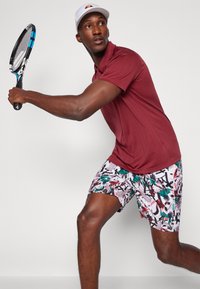 Bordo polo majica i hlače s uzorkom u bijelim, zelenim i crvenim nijansama. Drži plavi teniski reket, nosi bijelu kapu. Dinamičan položaj.