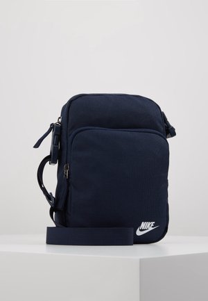 Kompaktní tmavě modrá crossbody taška Nike s předním zipovým kapsou a nastavitelným ramenním popruhem na bílém povrchu.