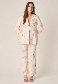 Woman wearing a cream floral suit with a long blazer and flared pants, standing with hands behind back against a plain background.