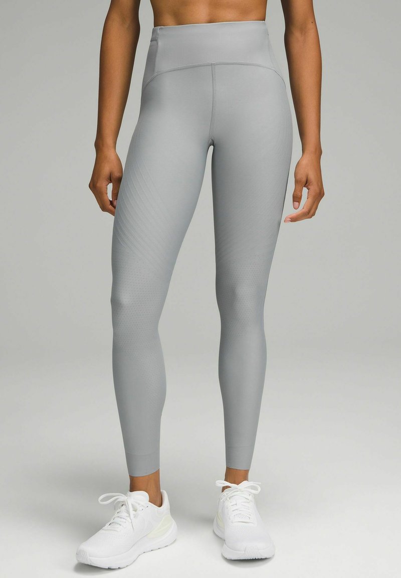 Legginsy sportowe o wysokim stanie, dopasowane, w jasnoszarym kolorze z teksturowanymi liniami. W połączeniu z białymi butami sportowymi, prezentującymi elegancki design.