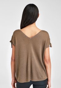 Mujer con cabello largo y oscuro, vistiendo una blusa marrón holgada de manga corta con un escote en forma de V en la parte de atrás y detalles de mangas atadas, de pie frente a un fondo liso.