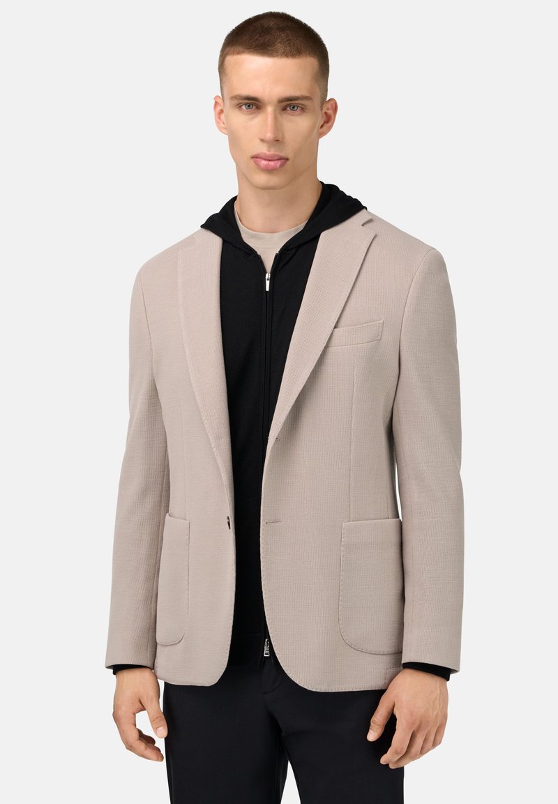 Ljusbeige texturerad blazer med två framfickor, bärs över en svart zip-up hoodie. Slim fit, enkelknäppning och noterade slag.