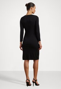 Zwart aansluitend jurk met lange mouwen, tot de knie, gladde textuur, sluiting met rits aan de achterkant, gedragen met zwarte hoge hakken sandalen.