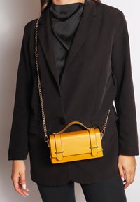 Borsa gialla in ecopelle di forma rettangolare, con due ganci metallici e una tracolla a catena. Indossata sopra un blazer nero.