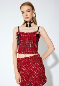 Punainen kudottu crop-top, jossa on franssit ja mustat hihnat, yhdistettynä vastaavaan hameeseen. Sisältää helmet ja nauhat koristuksina olkapäillä ja kaulalla.