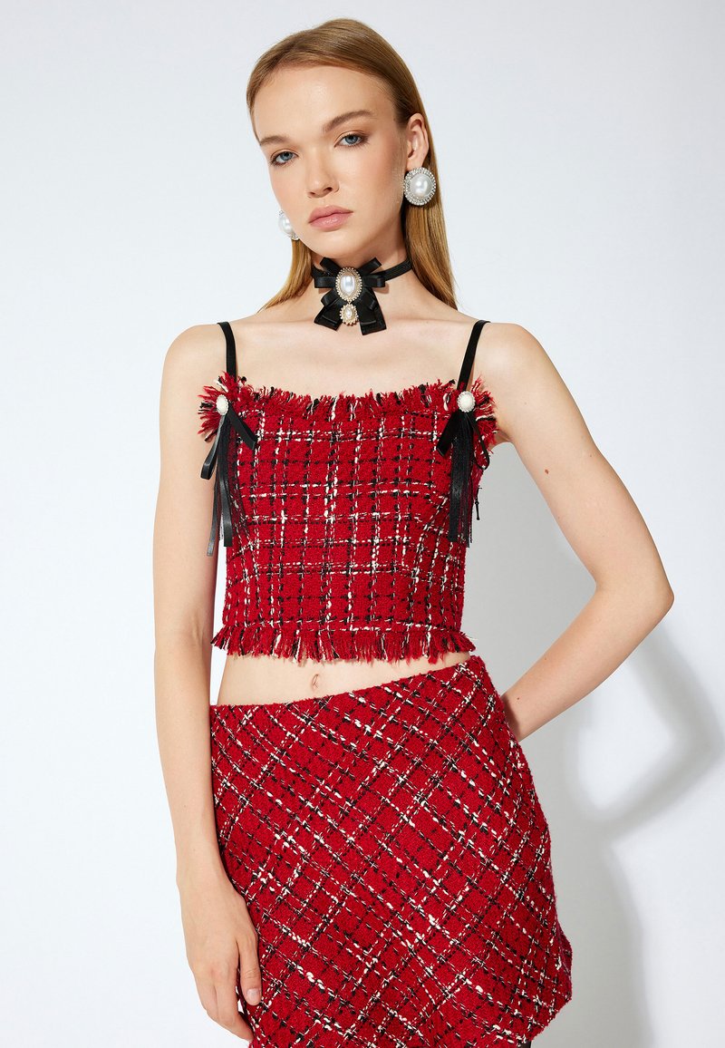 Punainen kudottu crop-top, jossa on franssit ja mustat hihnat, yhdistettynä vastaavaan hameeseen. Sisältää helmet ja nauhat koristuksina olkapäillä ja kaulalla.