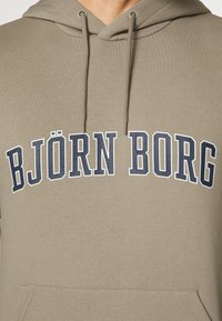 Khaki mikina s kapsou typu klokanka na přední straně, s výrazným textem "BJÖRN BORG" v námořnické modři a bílé barvě přes hruď, vyrobena z hladkého materiálu.