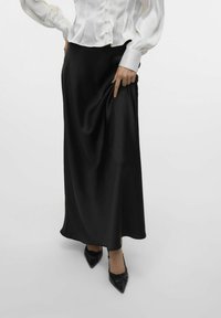 Zwarte satijnen maxi rok met een soepele textuur, voorzien van een lichte glans, gestyled met een witte blouse en puntige zwarte schoenen.