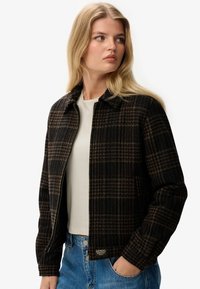 Zwarte en bruine geruite jas gemaakt van textuurstof met een kraag en ritssluiting, gedragen over een lichtgekleurde top.