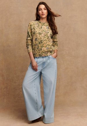 Donna in piedi con una mano in tasca, che indossa un maglione floreale e jeans a palazzo blu chiaro, contro uno sfondo beige semplice.