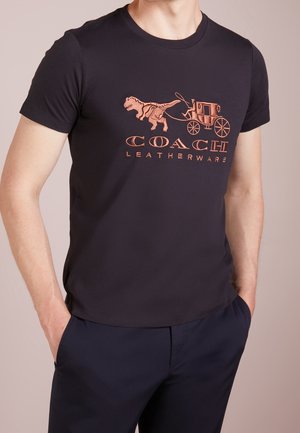 T-shirt noir à col rond avec un graphisme cuivre représentant un dinosaure et une calèche, accompagné du texte "COACH LEATHERWARE" en dessous. Matière en coton doux.