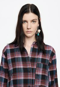 Camisa de cuadros en tonos de negro, rojo y azul. Hecha de una tela ligera con un frente abotonado y un bolsillo en el pecho. Cuenta con mangas largas.