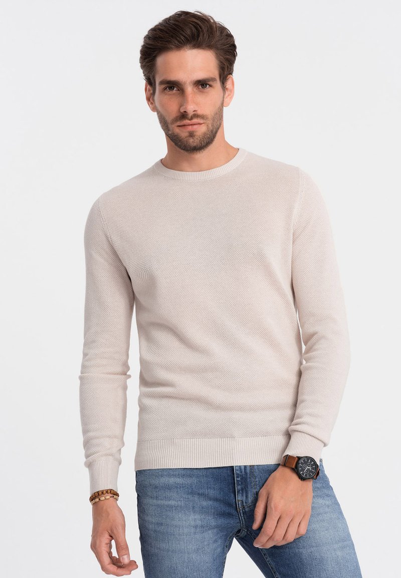 Maglione a maglia beige chiaro con scollo rotondo e maniche lunghe. Presenta polsini e orlo a costine. Indossato con jeans di denim e un orologio da polso.