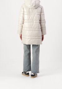 Světlý šedý nadýchaný kabát s kapucí, zdobený horizontálními švy a hladkou texturou, doplněný širokými džíny a černými teniskami.