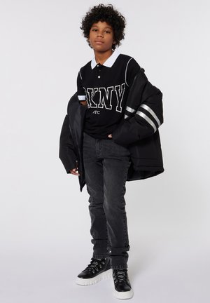 Polo noir avec col blanc, texte 'DKNY' en grand imprimé, associé à une veste matelassée noire avec des bandes argentées, un jean gris et des baskets noires.