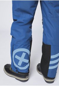 Blue waterproof pants feature a black textured panel at the ankle, adorned with a blue circular logo and striped accent. Black waterproof boots complete the look.