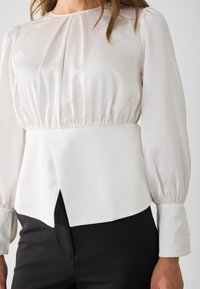 Hvit satengbluse med puffede lange ermer, rynket liv og peplum-kant. Har rund halslinning og knappede mansjetter.
