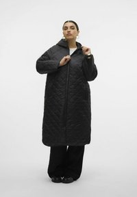 Gequilted zwarte lange jas met een capuchon, voorzien van een volledige ritssluiting en brede mouwen. Draagt zwarte broek en schoenen.