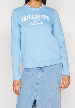 Personne portant un sweat Hollister California bleu clair et une jupe en jean bleu clair assortie avec une fente à l'avant.