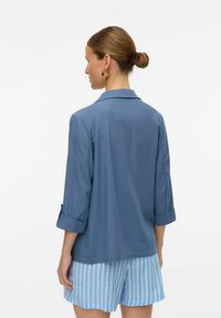 Šviesiai mėlyna, laisvai krentanti palaidinė su įpjovimu ant apykaklės ir sukišamomis rankovėmis, pagaminta iš lygaus audinio, derinama su dryžuotomis mėlynomis šortais.