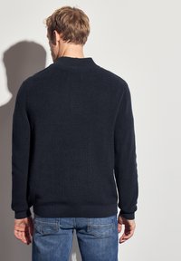Pull en tricot navy avec une texture côtelée, un col rond et des manches longues. Porté avec un jean bleu, mettant en avant une coupe ajustée.