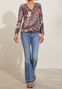 Kvinna som bär en långärmad blus med flerfärgat mönster, ljusblå flare-jeans och svarta sandaler med remmar.