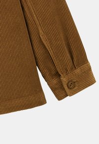 Manche de veste en velours côtelé marron, avec des nervures verticales, un bouton au poignet et une finition soignée le long de l'ourlet.