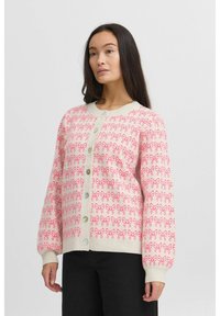Cardigan rose clair avec un motif de nœuds répétitif, des poignets côtelés, un col rond et une fermeture à boutons à l'avant avec de gros boutons.