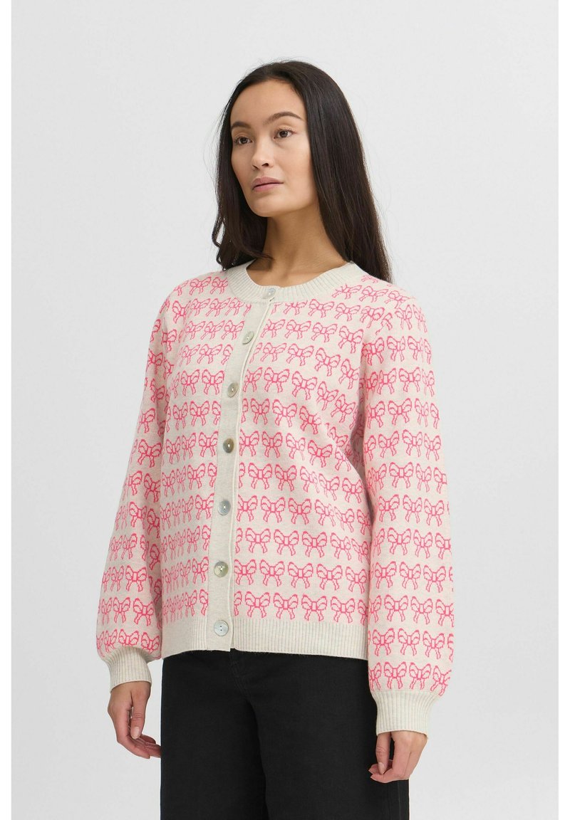 Cardigan rose clair avec un motif de nœuds répétitif, des poignets côtelés, un col rond et une fermeture à boutons à l'avant avec de gros boutons.