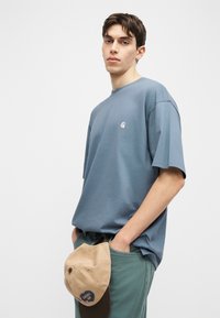 Νεαρός άνδρας που φοράει φαρδιά μπλε μπλούζα και πράσινο παντελόνι, κρατώντας ένα μπεζ καπέλο με μπλε λογότυπο στο δεξί του χέρι.