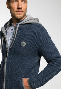 Donkerblauwe gebreide hoodie met grijze accenten, ritsluiting en een ronde logo patch op de borst. Gestructureerde stof en zijzakken.