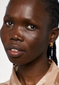 Primer plano de una persona de piel oscura con el cabello trenzado, que lleva una camisa beige clara y pequeños pendientes de aro dorados, mostrando una piel suave y una expresión natural.