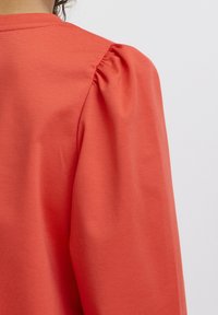 Indumento rosso a maniche lunghe con scollatura arrotondata e dettagli con maniche a sbuffo. Il tessuto appare liscio con una leggera trama.