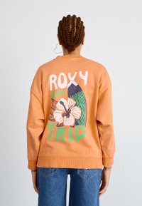Sweatshirt orange à col rond avec un design graphique représentant une planche de surf, une fleur et le texte "ROXY SURF TRIP" en vert et blanc. Matériel en coton, coupe décontractée.