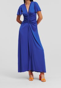 Kvinna iklädd en kungligt blå rynkad maxiklänning med korta ärmar, kombinerad med klarröda sandaler med öppen tå och blockklackar, som står mot en enfärgad bakgrund.