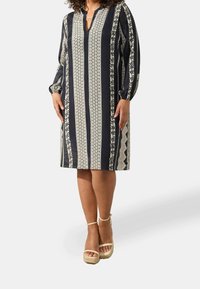 Tummansininen mekko, jossa on kukkakuvioita ja raidallista kuosia, kolmivartiset hihat ja lovikaulus. Yhdistetty beigeihin platform-sandaaleihin.