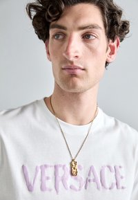 T-shirt bianco con logo "VERSACE" in viola, accompagnato da una collana con pendente dorato con dettagli intricati, indossata da un modello.