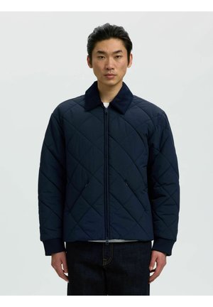 Jeune homme portant une veste matelassée bleu marine avec une fermeture éclair sur le devant et un pantalon foncé, debout sur un fond clair uni.