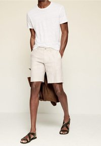 Weißes T-Shirt mit kurzen Ärmeln, beigefarbene Leinen-Shorts, braune Jacke übergeworfen, grüne Sandalen mit Kreuzriemen, die ein lässiges Sommeroutfit zeigen.