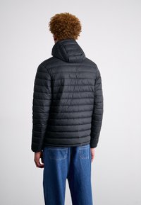 Czarna pikowana kurtka z kapturem, z poziomym przeszyciem, widok z tyłu, noszona z niebieskimi dżinsami. Gładka faktura, lekki materiał.