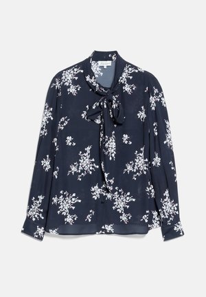 Vrouw draagt een zwarte blouse met bloemenprint en een strik, zwarte op maat gemaakte broek, zwarte hoge hakken en houdt een kleine zwarte clutch vast.