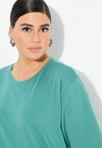 Türkise T-Shirts mit Rundhalsausschnitt und kurzen Ärmeln, aus einem glatten Stoff gefertigt. Das Modell trägt große silberne Kreolen.